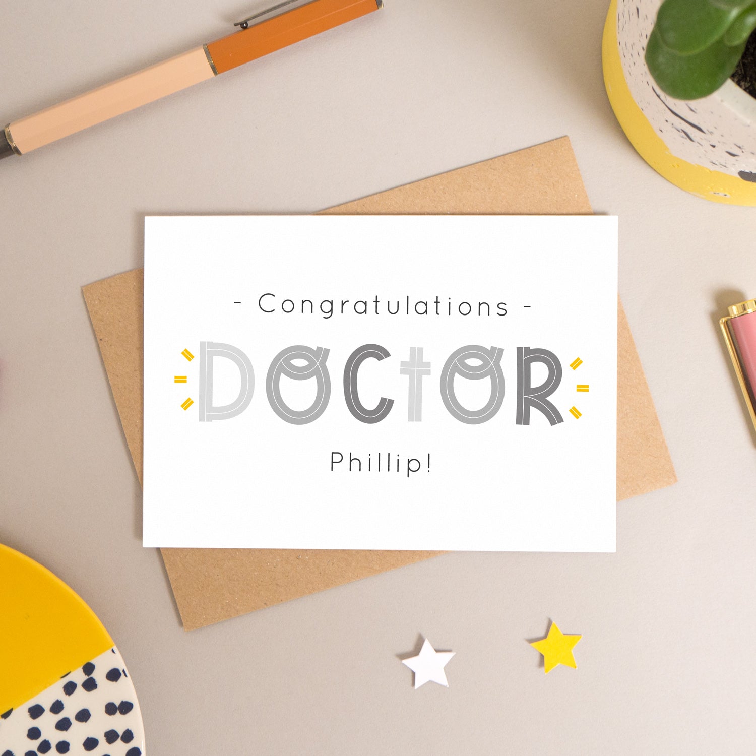 This personalised doctor card has been shot flatlay style looking directly over the top of the card. It is lying flat on it’s kraft brown envelope, on a warm grey background. Surrounding the card are pens, a clip, a plant and a trinket dish. This version of the card is in varying tones of grey.