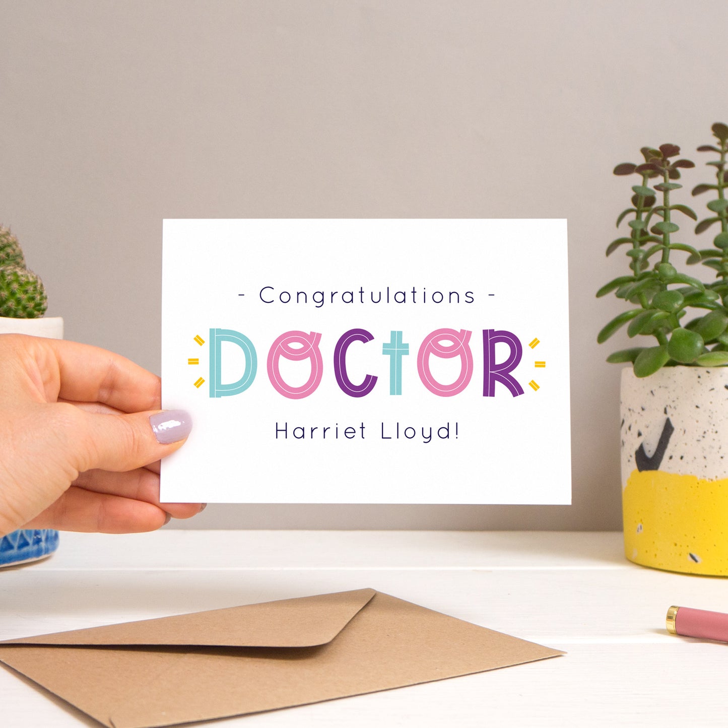 A personalised doctor card held over a warm grey and white background with potted plants peeping the sides. Behind the card is a kraft brown envelope that comes with the card. The text on this version of the card is in varying tones of navy and pink, purple and blue.