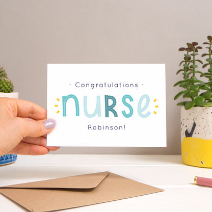 A personalised nurse card held over a warm grey and white background with potted plants peeping the sides. Behind the card is a kraft brown envelope that comes with the card. The text on this version of the card is in varying tones of navy and blue.