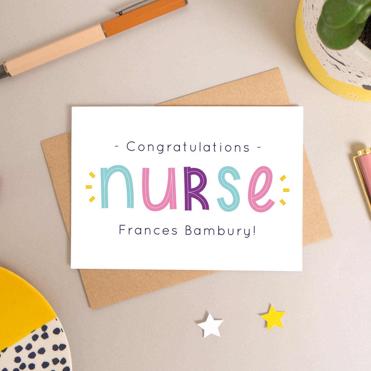 This personalised nurse card has been shot flatlay style looking directly over the top of the card. It is lying flat on it’s kraft brown envelope, on a warm grey background. Surrounding the card are pens, a clip, a plant and a trinket dish. This version of the card is in varying tones of navy and pink, purple and blue.