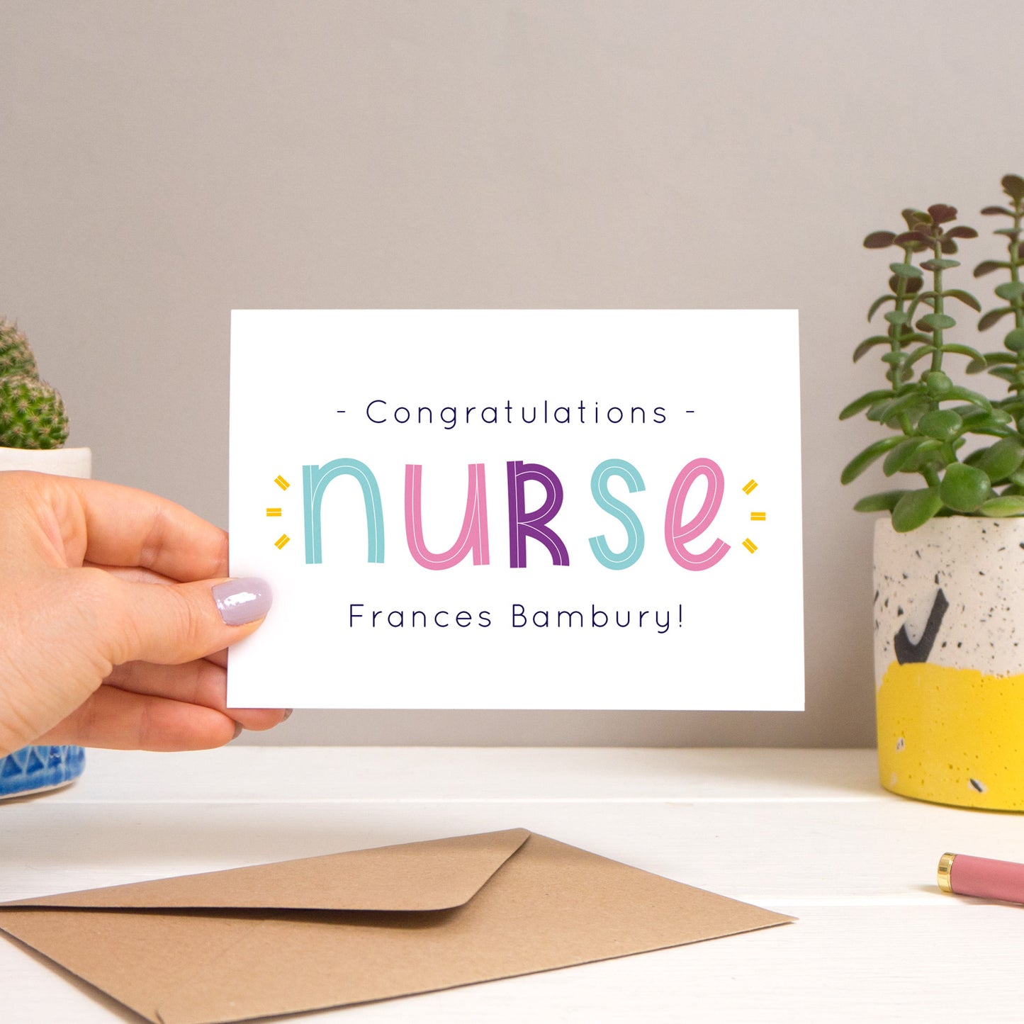 A personalised nurse card held over a warm grey and white background with potted plants peeping the sides. Behind the card is a kraft brown envelope that comes with the card. The text on this version of the card is in varying tones of navy and pink, purple and blue.