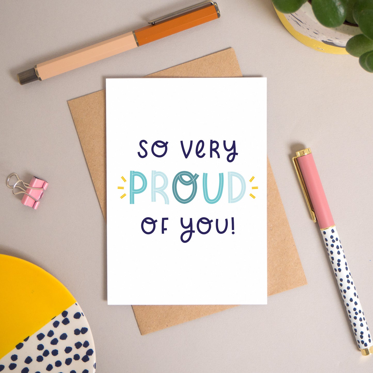 This ‘so very proud of you’ card has been shot flatlay style looking directly over the top of the card. It is lying flat on it’s kraft brown envelope, on a warm grey background. Surrounding the card are pens, a clip, a plant and a trinket dish. This version of the card is in varying tones of navy and blue.