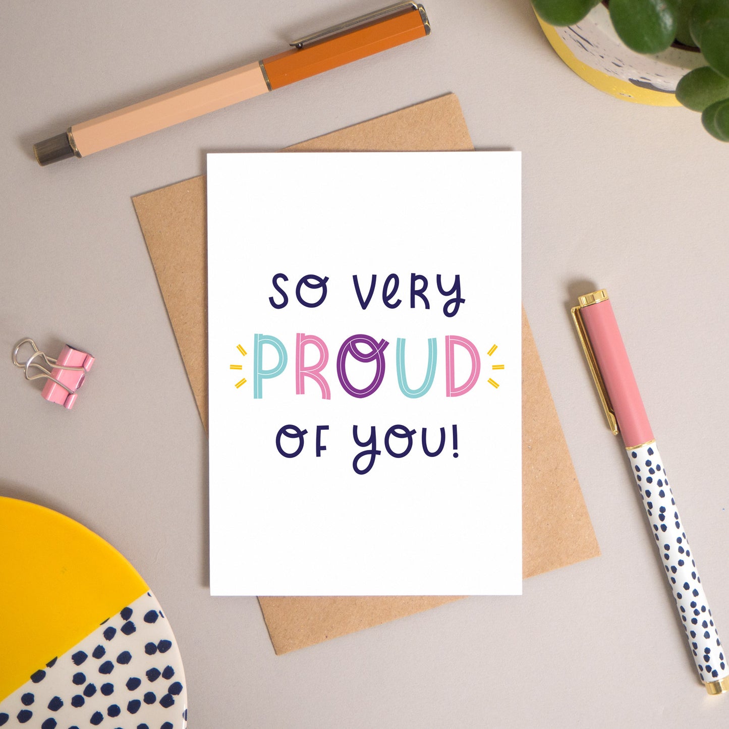 This ‘so very proud of you’ card has been shot flatlay style looking directly over the top of the card. It is lying flat on it’s kraft brown envelope, on a warm grey background. Surrounding the card are pens, a clip, a plant and a trinket dish. This version of the card is in varying tones of navy and pink, purple and blue.
