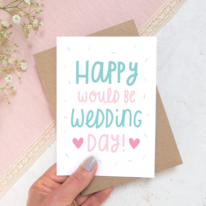 Happy would be wedding day card in teal and pink photographed in a hand against a pink, and white background with a hint of flowers.