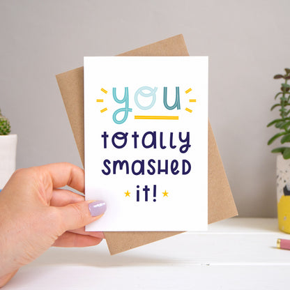 A ‘you totally smashed it’ card held over a warm grey and white background with potted plants peeping the sides. Behind the card is a kraft brown envelope that comes with the card. The text on this version of the card is in varying tones of navy and blue.