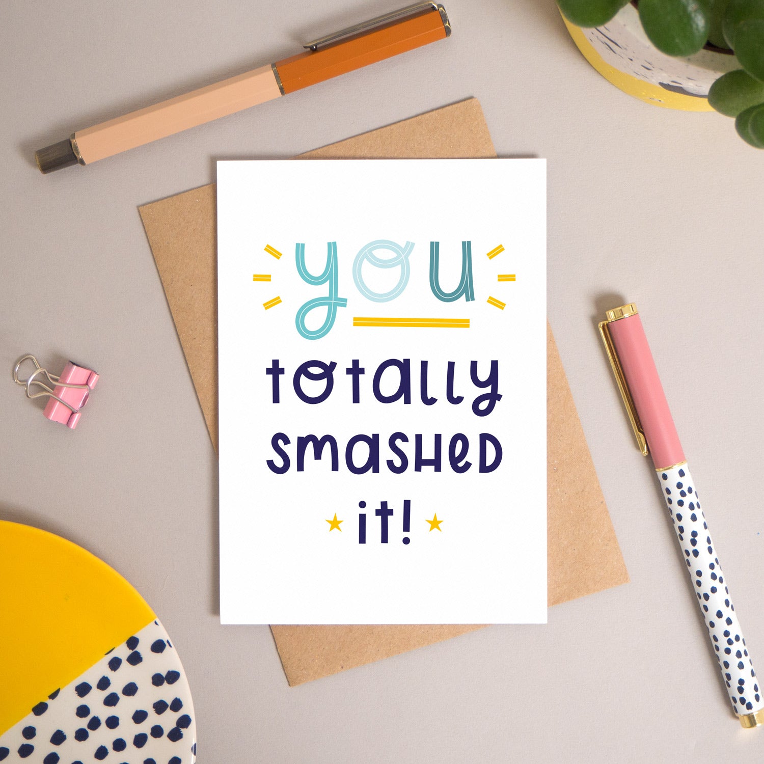 This ‘you totally smashed it’ card has been shot flatlay style looking directly over the top of the card. It is lying flat on it’s kraft brown envelope, on a warm grey background. Surrounding the card are pens, a clip, a plant and a trinket dish. This version of the card is in varying tones of navy and blue.