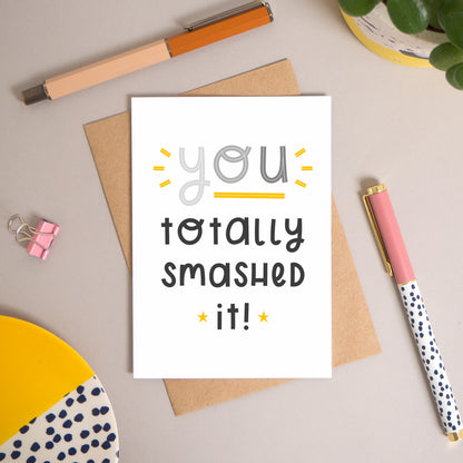 This ‘you totally smashed it’ card has been shot flatlay style looking directly over the top of the card. It is lying flat on it’s kraft brown envelope, on a warm grey background. Surrounding the card are pens, a clip, a plant and a trinket dish. This version of the card is in varying tones of grey.