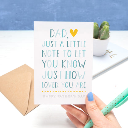 A father's day card by Joanne Hawker being held in her right hand, with a green pen on a white and blue background. The card reads "Dad, just a little note to let you know just how loved you are - happy fathers day". The letters are in varying shades of blue with a yellow heart.