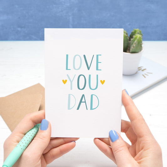 Love you dad card held over a white and blue background by Joanne Hawker in her somerset studio. The text on the card is in varying tones of blue with two yellow hearts surrounding the word 'you'.