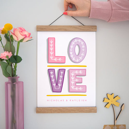 A personalised 'love' print featuring hand drawn typography in varying shades of pink and purple with space for the names of the couple. The image has a vase of flowers to the left, the print being held centre and a small wooden flower in the bottom right set on a grey background.
