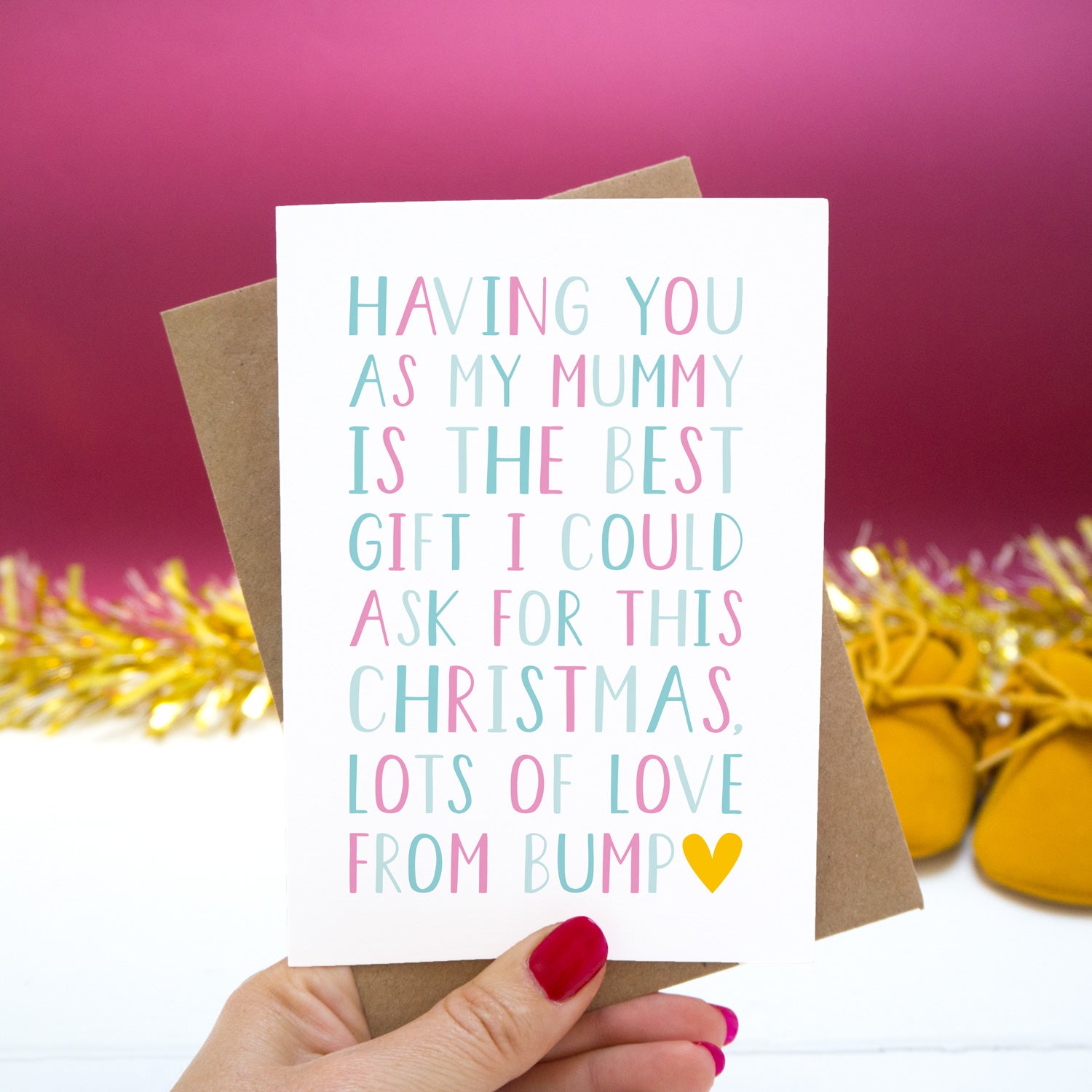 Having you as my Mummy is the best gift I could ask for this Christmas, lots of love from bump." - Christmas bump card with pink and blue text set on a red background with gold tinsel and mustard shoes.