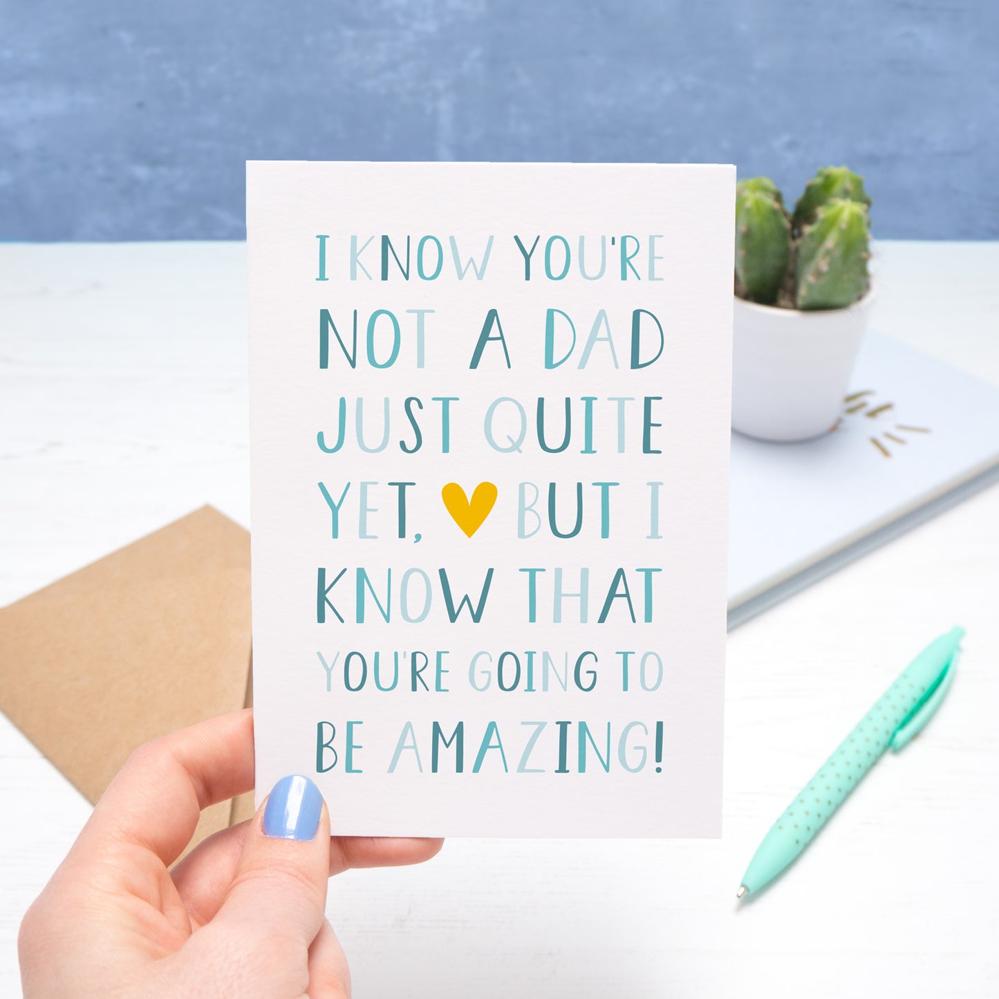 A typographic father's day card by Joanne Hawker, held on a white and blue background. The card reads "I know you're not a dad just quite yet but I know that you're going to be amazing". The letters are in varying tones of blue with a yellow heart near the centre of the card!