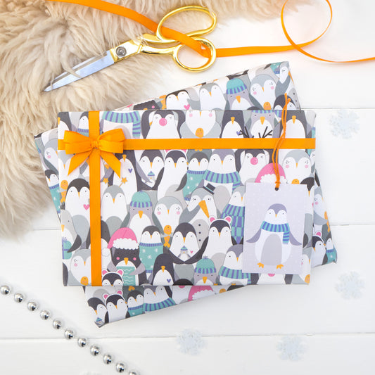 A parade of penguins wearing a variety of Christmas fancy dress printed onto white gift wrap. Matching penguin tag shown on the bottom right of the wrapping paper. The present is strung with orange ribbon and features a small bow.