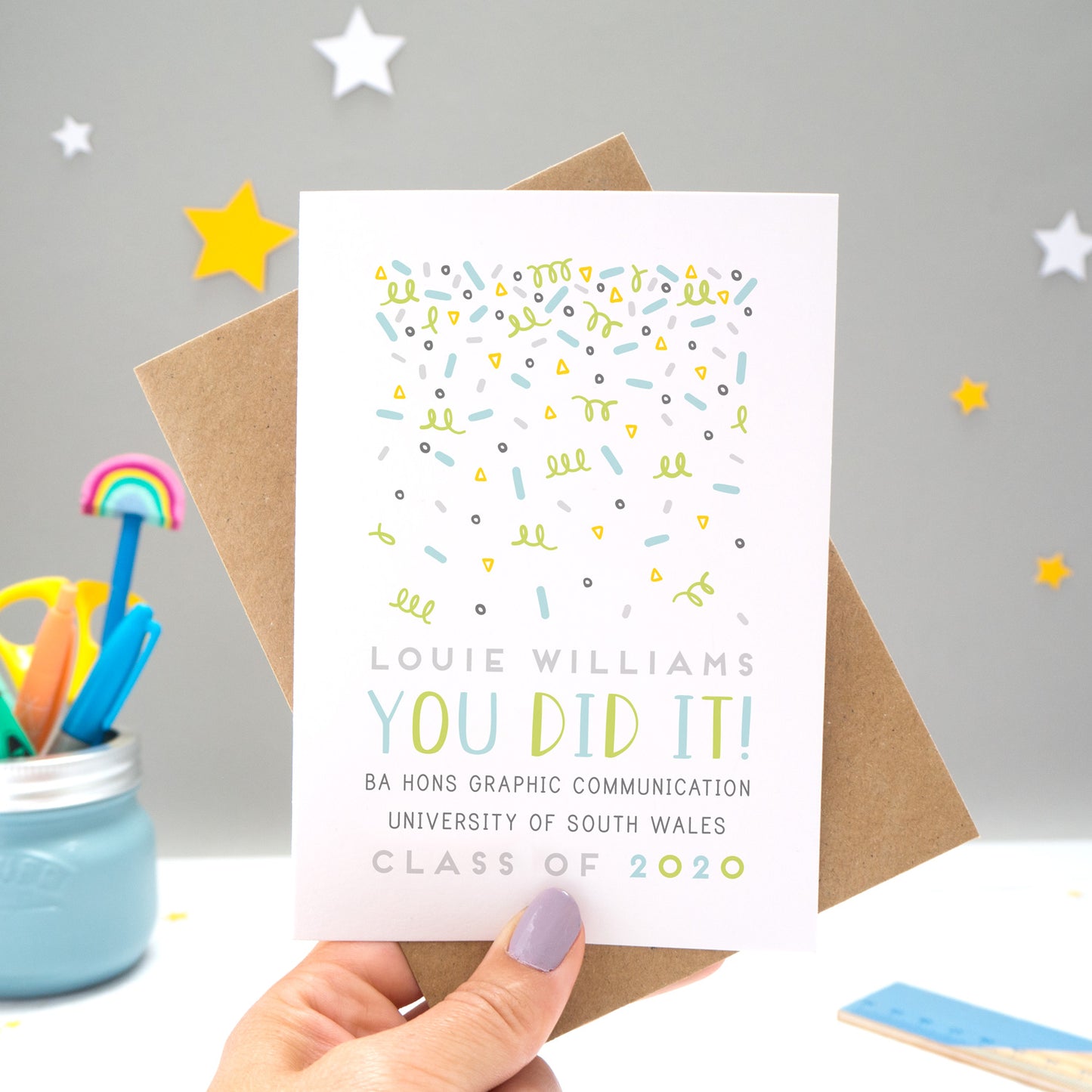 A personalised graduation card designed and made by Joanne Hawker in her somerset studio being held against a kraft brown envelope over a grey background with yellow and white stars. The confetti illustration and text is in varying tones of grey, blue and green.