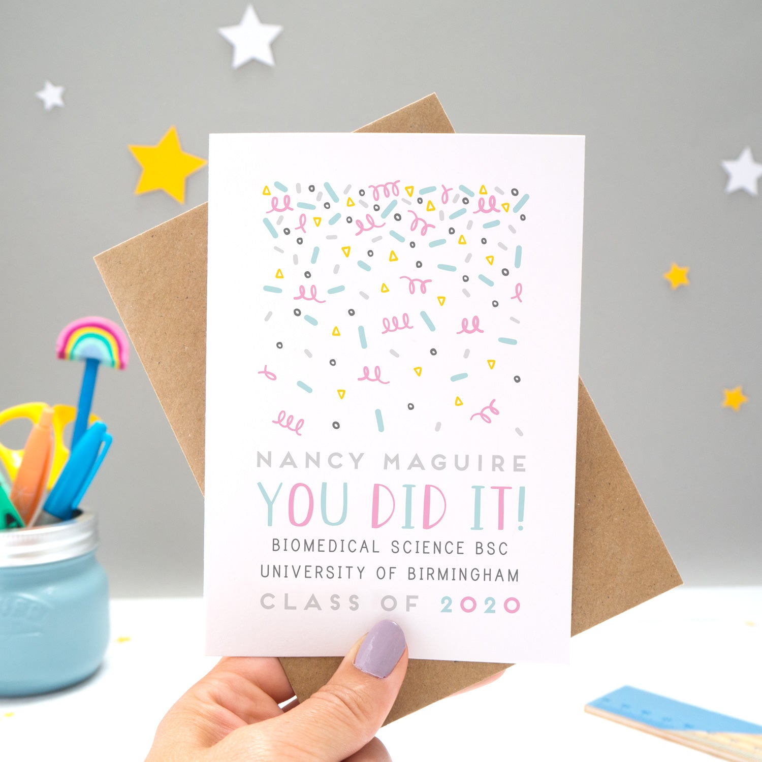 A personalised graduation card designed and made by Joanne Hawker in her somerset studio being held against a kraft brown envelope over a grey background with yellow and white stars. The confetti illustration and text is in varying tones of grey, blue and pink.
