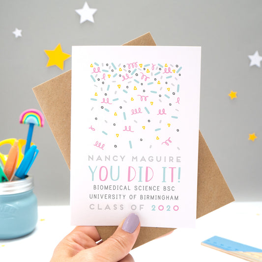 A personalised graduation card designed and made by Joanne Hawker in her somerset studio being held against a kraft brown envelope over a grey background with yellow and white stars. The confetti illustration and text is in varying tones of grey, blue and pink.