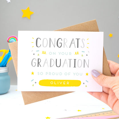 This personalised graduation card reads 'congrats on your graduation so proud of you [insert name].' The card is being held by Joanne Hawker in her somerset studio against a grey background with a kraft brown envelope with yellow and white stars. The card features grey text with varying tones of green, blue and yellow.