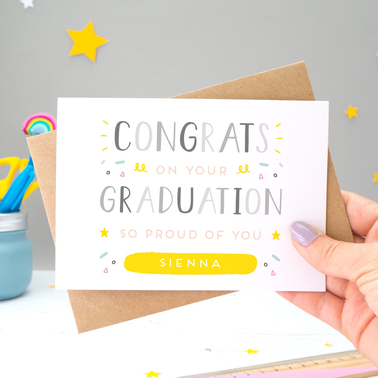 This personalised graduation card reads 'congrats on your graduation so proud of you [insert name].' The card is being held by Joanne Hawker in her somerset studio against a grey background with a kraft brown envelope with yellow and white stars. The card features grey text with varying tones of pink, blue and yellow.