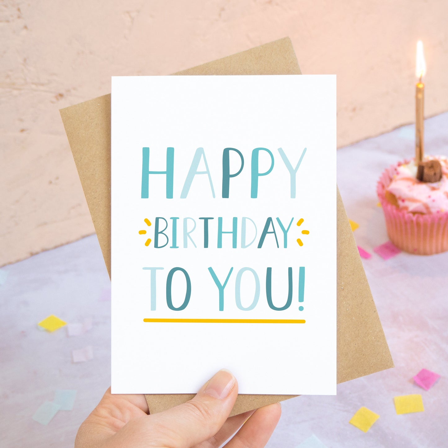 A happy birthday to you card in blue photographed over a grey and peach background with pieces of confetti and a pink cupcake with a single candle.