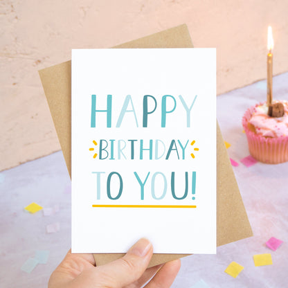 A happy birthday to you card in blue photographed over a grey and peach background with pieces of confetti and a pink cupcake with a single candle.