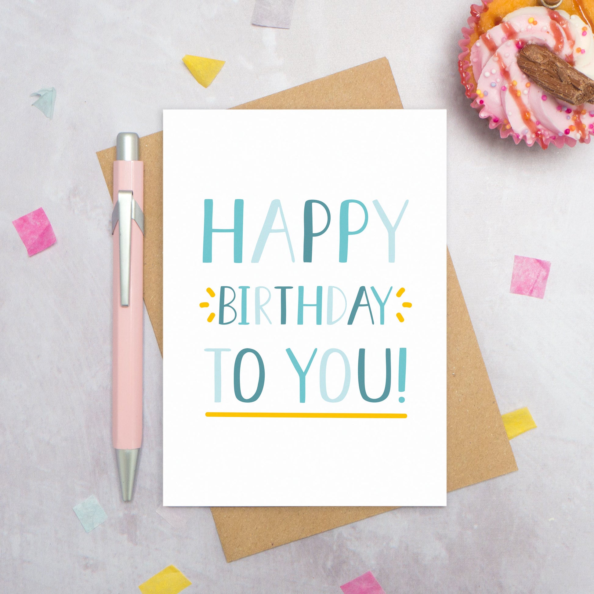 A happy birthday to you card photographed from directly above. The card is lying on its kraft brown envelope which is on a grey surface. There is also a pink pen, confetti and a pink cupcake! This is the blue colour palette!