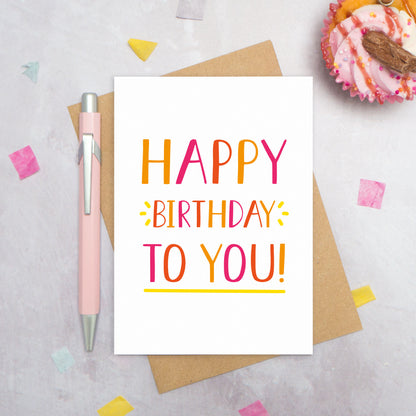 A happy birthday to you card photographed from directly above. The card is lying on its kraft brown envelope which is on a grey surface. There is also a pink pen, confetti and a pink cupcake! This is the orange colour palette!