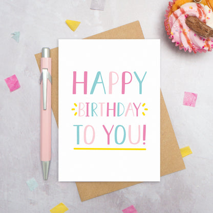 A happy birthday to you card photographed from directly above. The card is lying on its kraft brown envelope which is on a grey surface. There is also a pink pen, confetti and a pink cupcake! This is the pink and blue colour palette!