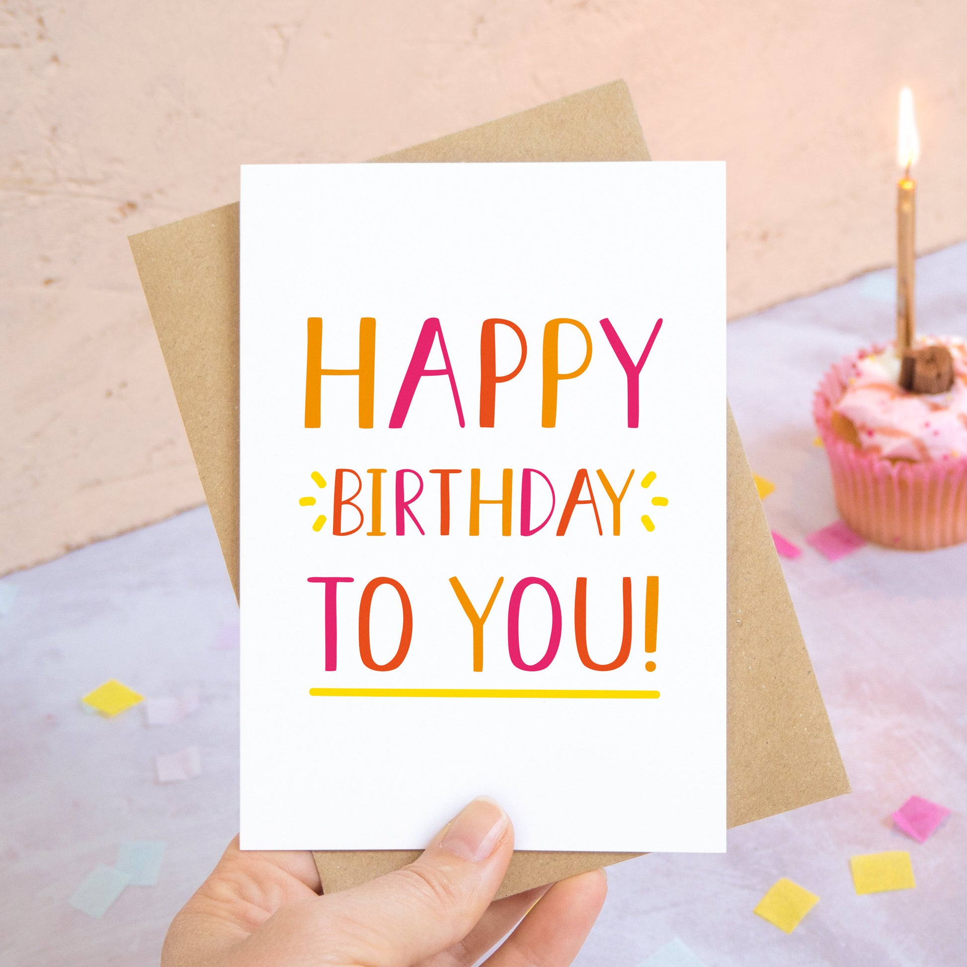 A happy birthday to you card in orange photographed over a grey and peach background with pieces of confetti and a pink cupcake with a single candle.