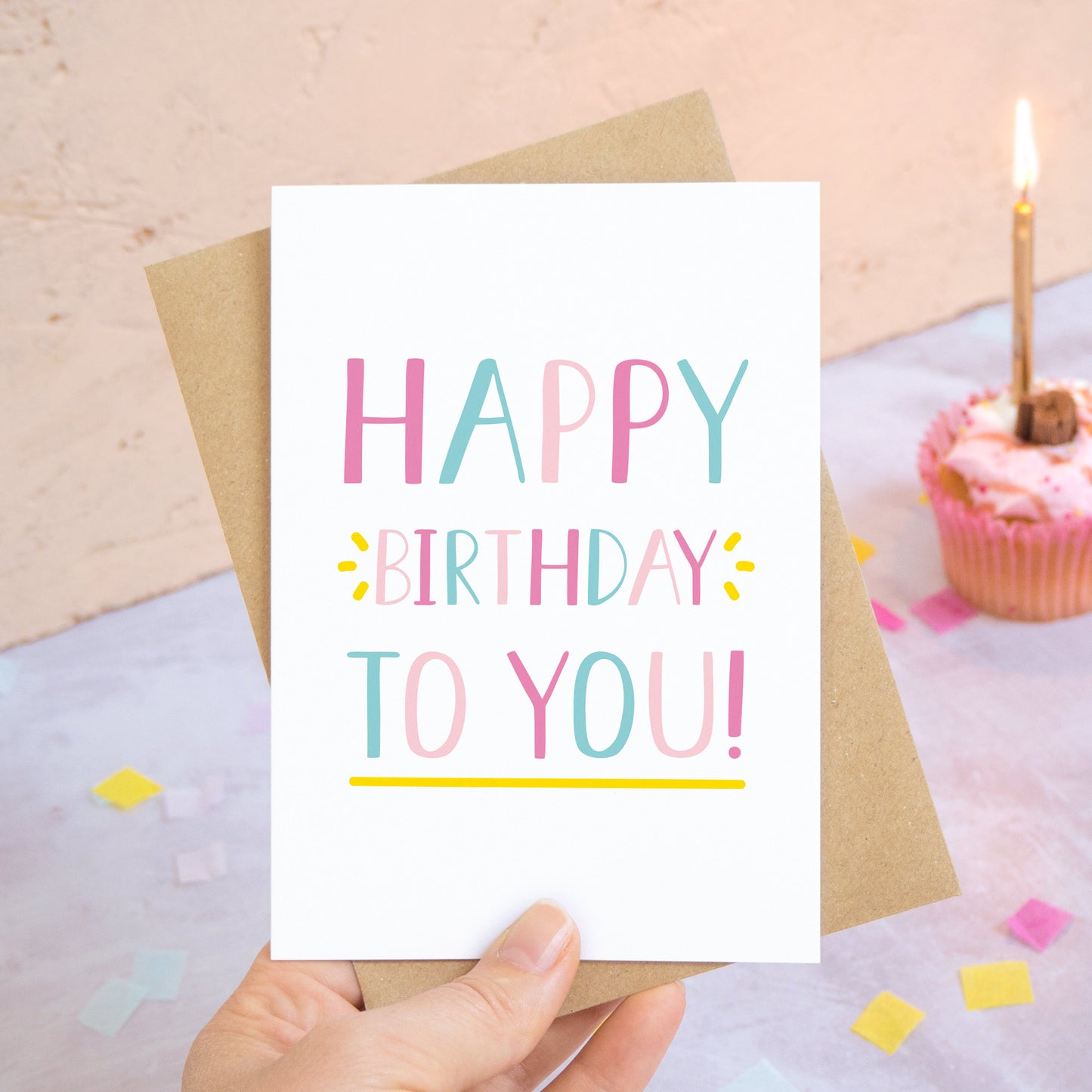 A happy birthday to you card in pink and blue photographed over a grey and peach background with pieces of confetti and a pink cupcake with a single candle.